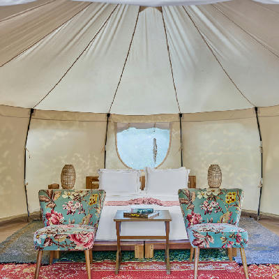 Interior of a round glamping tent with a white bed, floral armchairs, and woven rugs.