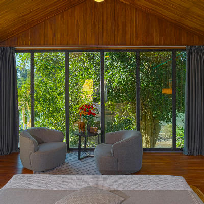 Two grey armchairs face floor-to-ceiling glass windows overlooking dense green jungle foliage.