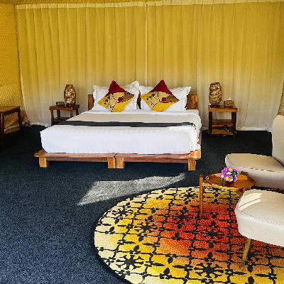 A glamping tent bedroom with a white-dressed double bed, colourful patterned rug, and warm yellow canvas walls.