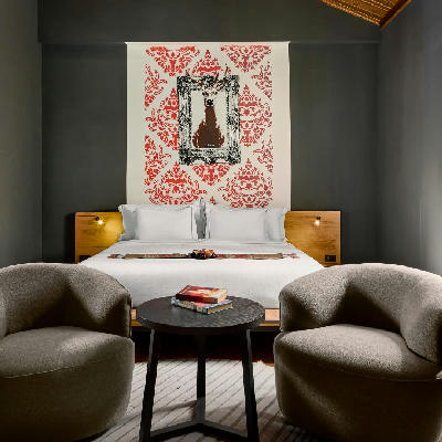 A hotel bedroom with a red patterned wall textile above a white-dressed double bed flanked by teak nightstands and two grey armchairs.