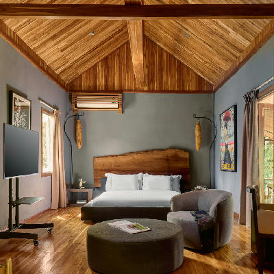 Spacious hotel suite with vaulted teak ceiling, live-edge wooden headboard, and muted grey walls.