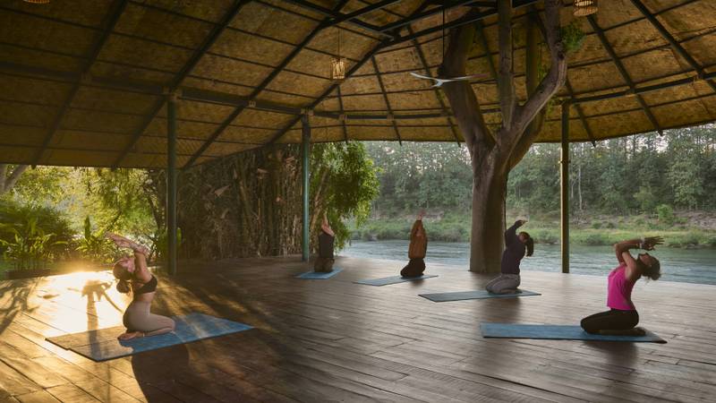 Three guests practising yoga on mats inside an open teak pavilion overlooking a river.