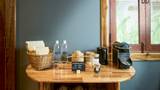 In-room amenity station with glass water bottles, rattan basket, coffee machine, and tea on a teak dresser.