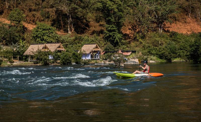 Guest kayaking through gentle rapids on Nam Khan River with traditional thatched riverside villas and lush hillside forest backdrop