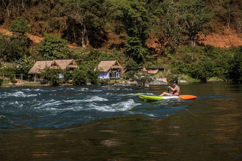 Guest kayaking through gentle rapids on Nam Khan River with traditional thatched riverside villas and lush hillside forest backdrop