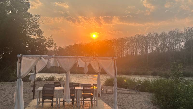 White canopy dining setup on sandy riverbank at sunset, with table and chairs facing the Nam Khan River through dramatic orange skies
