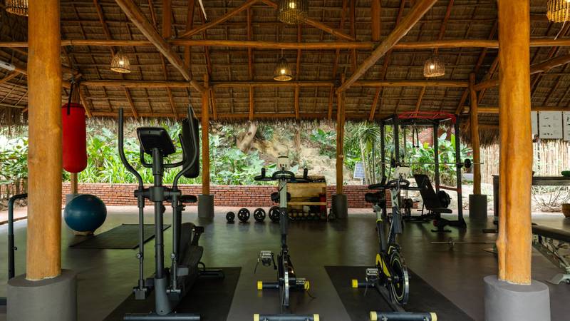 Open-air fitness center with cardio equipment, weights, and spin bikes under traditional thatched roof surrounded by tropical gardens
