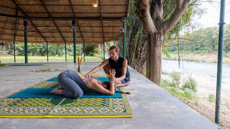 Yoga instructor guiding guest in riverside pavilion at The Namkhan resort, Luang Prabang, with natural teak structure and river views