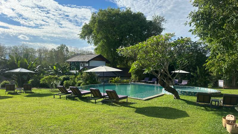 Turquoise swimming pool surrounded by manicured lawn with teak loungers, white umbrellas, and tropical trees under blue sky at Namkhan