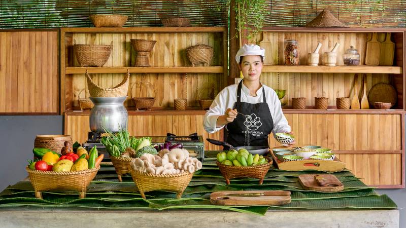 A chef in uniform stands behind rattan baskets of fresh vegetables and fruit in a wooden kitchen.