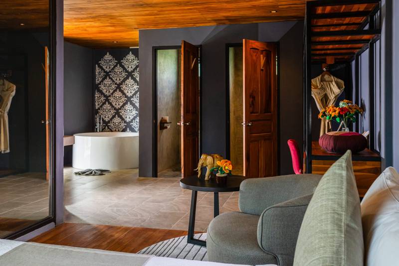 View into ensuite bathroom with freestanding tub and ornate wall art at The Namkhan resort suite in Luang Prabang, featuring warm wood tones and contemporary...