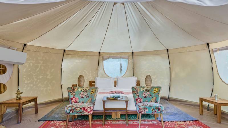 Interior of a round glamping tent with a white bed, floral armchairs, and woven rugs.