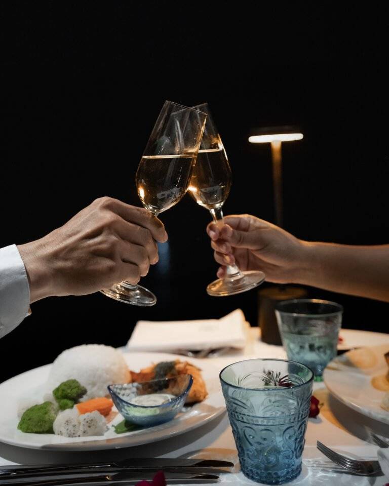 Couple toasting white wine glasses over fine dining plates at The Namkhan restaurant in Luang Prabang, with blue glassware and candlelight.