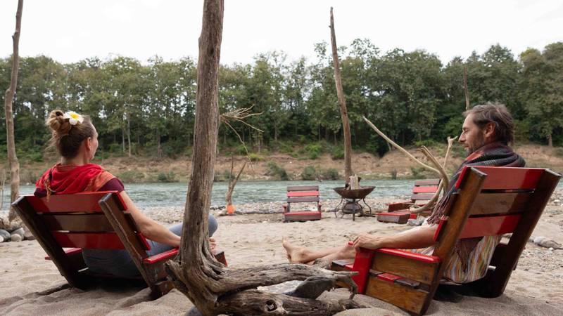 Two guests relaxing in wooden lounge chairs on a sandy riverbank with turquoise water, driftwood, and fire pit at Namkhan eco-lodge