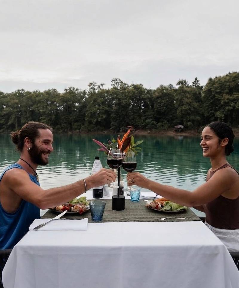 Couple raising wine glasses in a toast at an intimate riverside dining table with tropical flowers, surrounded by emerald waters and forest