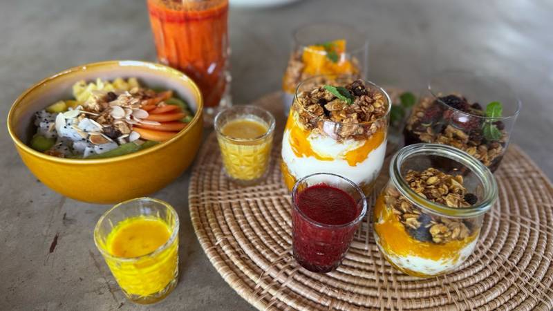 Colorful breakfast spread featuring acai bowl with tropical fruit, layered granola parfaits, fresh pressed juices on woven placemat