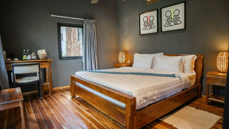 A king bed with white linens on a teak frame in a grey-walled room with artwork above.