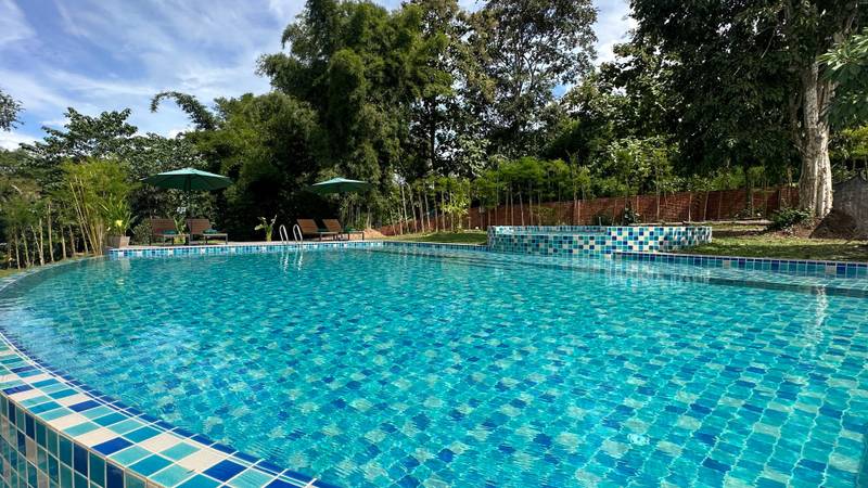 Large mosaic-tiled swimming pool with turquoise water surrounded by tropical trees, green umbrellas, and teak loungers at Namkhan resort