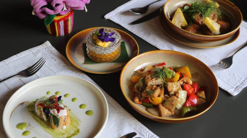 Four artfully plated Lao dishes including stir-fried vegetables, herb-crusted fish, and mango dessert served on golden-rimmed ceramic bowls — The Namkhan Luang Prabang