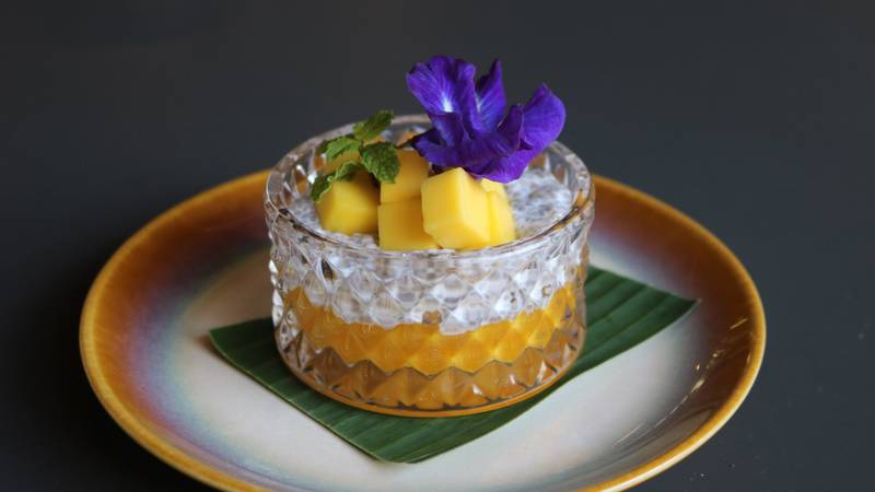 Crystal glass filled with golden mango sticky rice topped with fresh mango cubes, mint, and purple butterfly pea flower on banana leaf