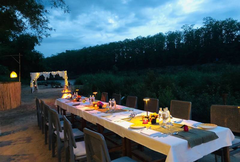 Long outdoor dining table set for evening meal beside the Nam Khan river, surrounded by lush jungle vegetation and ambient candlelight