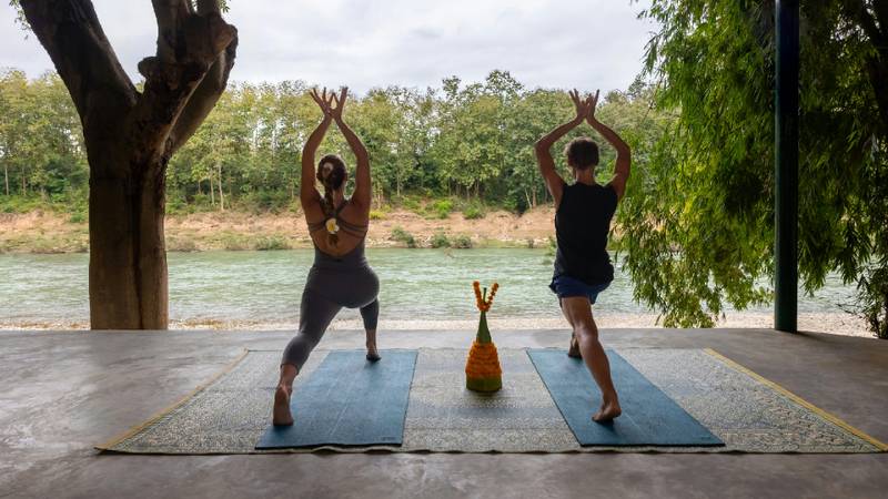 Two people practicing yoga on blue mats facing the Namkhan river with trees beyond.