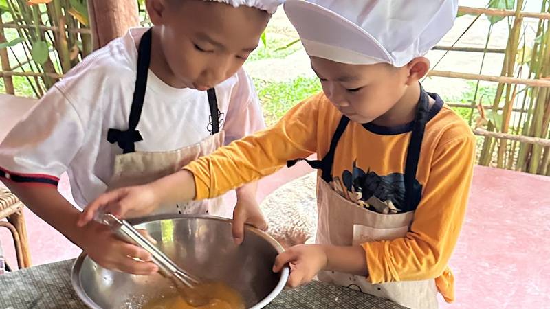 Two children in chef hats and aprons whisking ingredients in a metal bowl during a cooking class at an outdoor pavilion kitchen