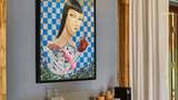 A bold figurative painting with a checkered background hangs above a timber-framed room shelf with wine glasses and accessories.