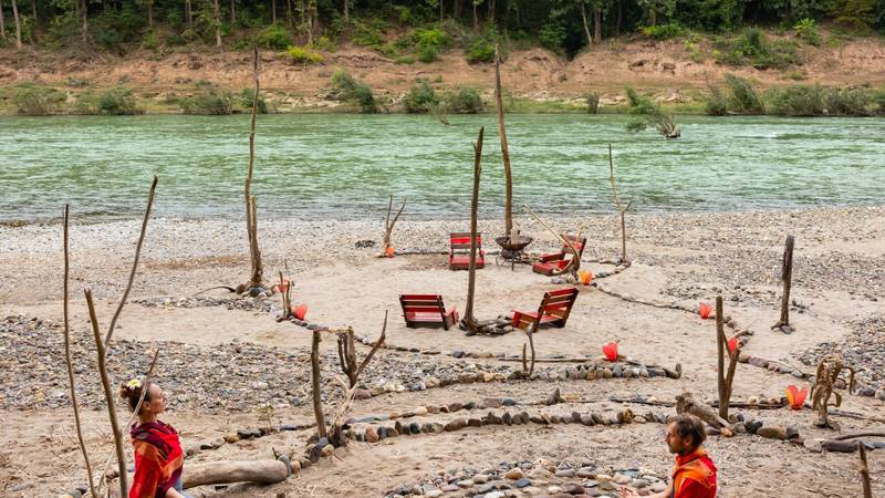 Two guests in traditional robes meditating in a stone circle labyrinth on the Nam Khan riverbank, surrounded by natural driftwood and red lounge chairs