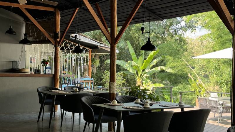 Open-air restaurant with wooden beams, black pendant lights, modern dark chairs set for breakfast overlooking lush tropical jungle