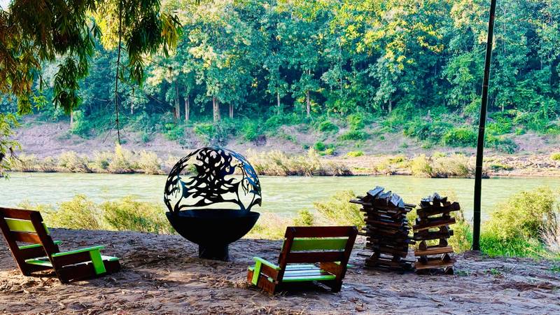 Decorative fire pit beside wooden lounge chairs overlooking the Nam Khan river, with lush forest backdrop and natural riverbank setting