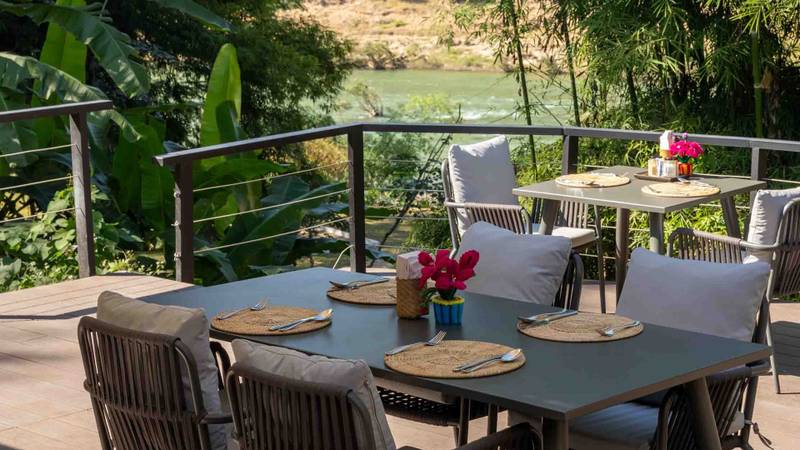 Private terrace dining tables set for breakfast overlooking the Namkhan River, framed by tropical bamboo and banana trees under shade umbrellas