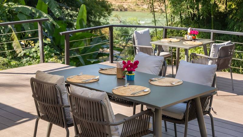 Outdoor terrace dining setup with river views at The Namkhan resort in Luang Prabang, featuring natural woven placemats and tropical foliage.