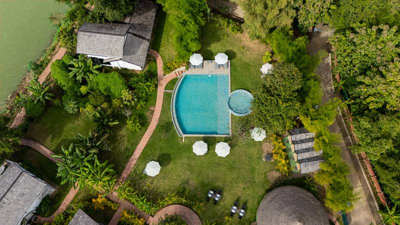 Aerial view of turquoise swimming pool surrounded by white umbrellas, lush tropical gardens, and traditional Lao buildings beside the Nam Khan River