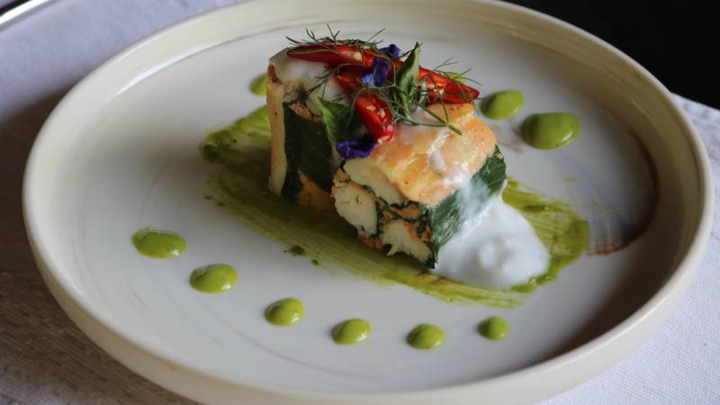 Seared fish fillet with crispy skin, garnished with fresh herbs, chili, and edible flowers on herb oil dots, served on cream ceramic plate — The Namkhan Luang Prabang
