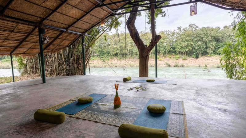 Open-air yoga pavilion with traditional bamboo roof overlooking the Nam Khan River, featuring meditation mats and cushions arranged around a central tree