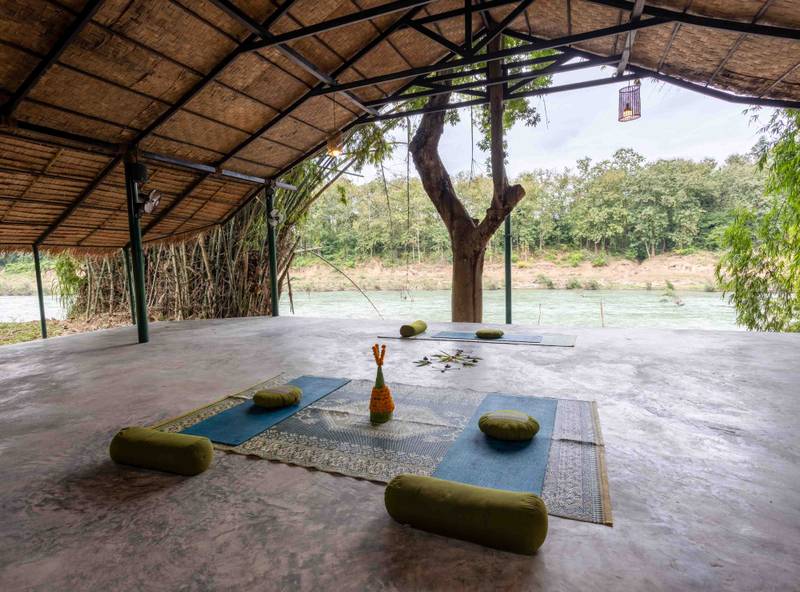 Open-air yoga pavilion with traditional bamboo roof overlooking the Nam Khan River, featuring meditation mats and cushions arranged around a central tree
