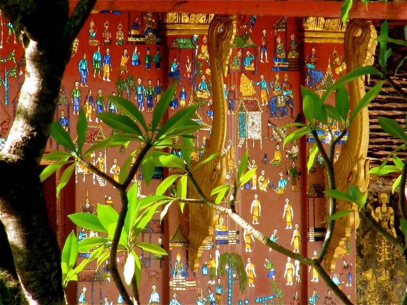 Vibrant traditional Lao temple mural with gold leaf details and colorful figures, framed by tropical foliage at The Namkhan in Luang Prabang, Laos.