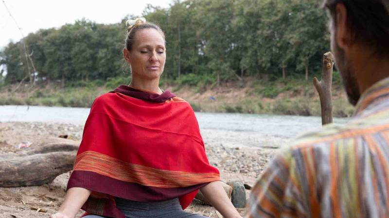 Two guests practicing guided meditation on smooth river stones beside the Nam Khan, surrounded by lush jungle and driftwood at Namkhan resort
