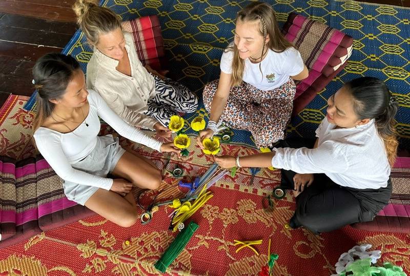 Four guests sitting cross-legged on traditional Lao textiles, crafting decorative items with yellow flowers during a cultural workshop