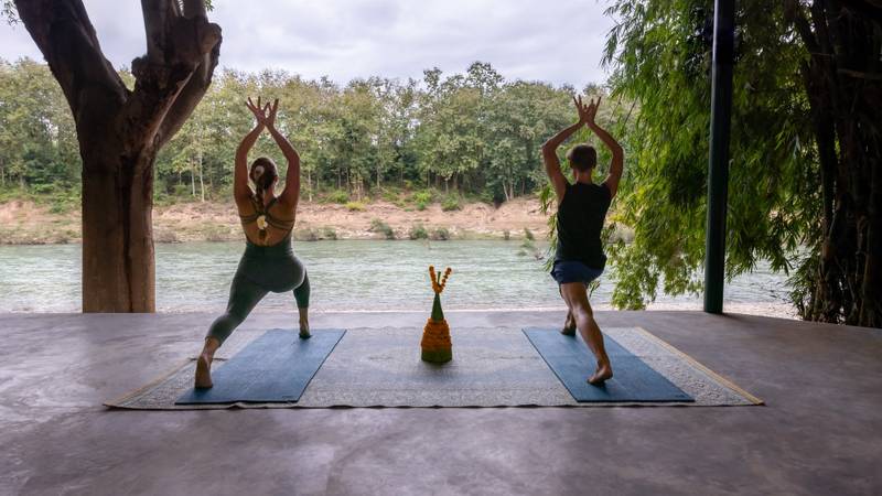 Two guests practicing yoga in warrior pose on mats at The Namkhan's riverside pavilion overlooking the Nam Khan River in Luang Prabang, Laos.