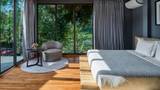 A villa bedroom with teak floors, grey armchair, and floor-to-ceiling jungle-view windows.
