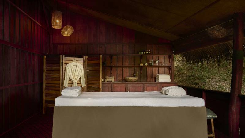 A spa treatment room with a massage table, white robe, and dark teak shelving.