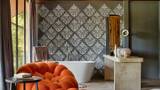 Freestanding white bathtub against a grey damask wall beside an orange sculptural armchair.