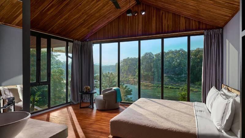 A sunlit villa bedroom with teak ceiling, floor-to-ceiling windows overlooking the Namkhan River and jungle.