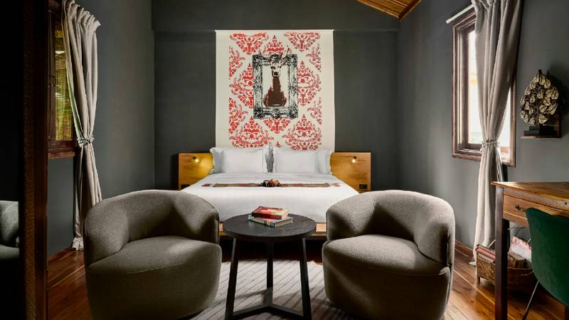 A hotel bedroom with a red patterned wall textile above a white-dressed double bed flanked by teak nightstands and two grey armchairs.