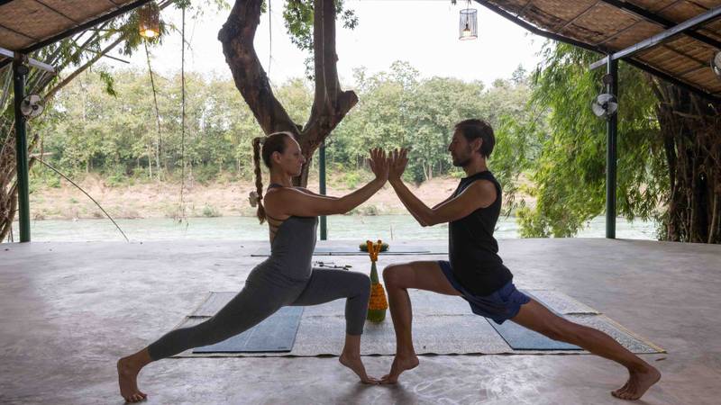 Two guests practice partner yoga in open-air bamboo pavilion overlooking the Nam Khan River, natural light filtering through woven roof