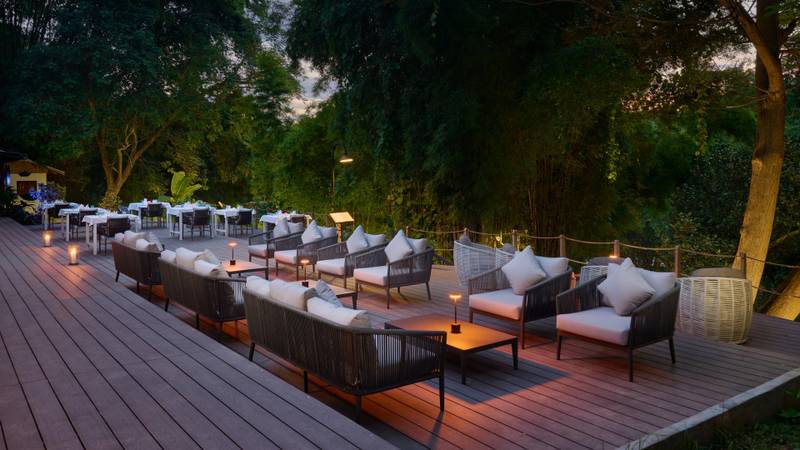 Wooden deck terrace with lounge seating and white-cloth dining tables surrounded by tropical forest at dusk, ambient lighting creates warm glow