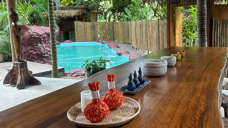 Traditional Lao herbal compress pouches and wellness oils arranged on natural wood table overlooking turquoise pool at The Namkhan spa in Luang Prabang
