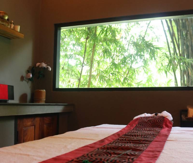 Spa treatment room at The Namkhan in Luang Prabang featuring massage bed with red silk runner and bamboo garden views through picture window